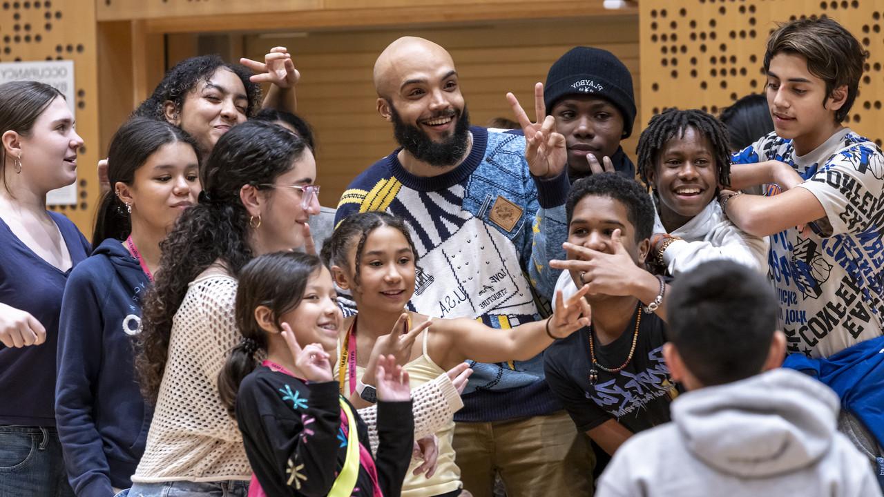 A joyful moment among young students gathered around Marcus Norris in an orchestral rehearsal studio at Juilliard. Norris is smiling broadly and wearing a stylish, patterned sweater. He stands at the center, surrounded by the enthusiastic students posing playfully—some making peace signs, while others smile or gesture animatedly towards the camera.