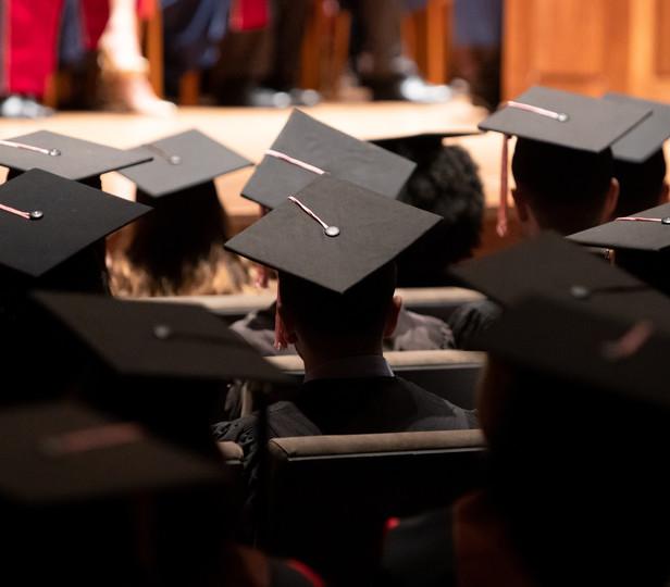 A group of graduates during a commencement ceremony. They are wearing traditional black caps and gowns. The setting is Alice Tully Hall with rows of seating filled with the graduates. The angle of the photo suggests it’s taken from the back of the hall, focusing on the back of the graduates' heads and their mortarboards, with one in the foreground serving as a focal point.